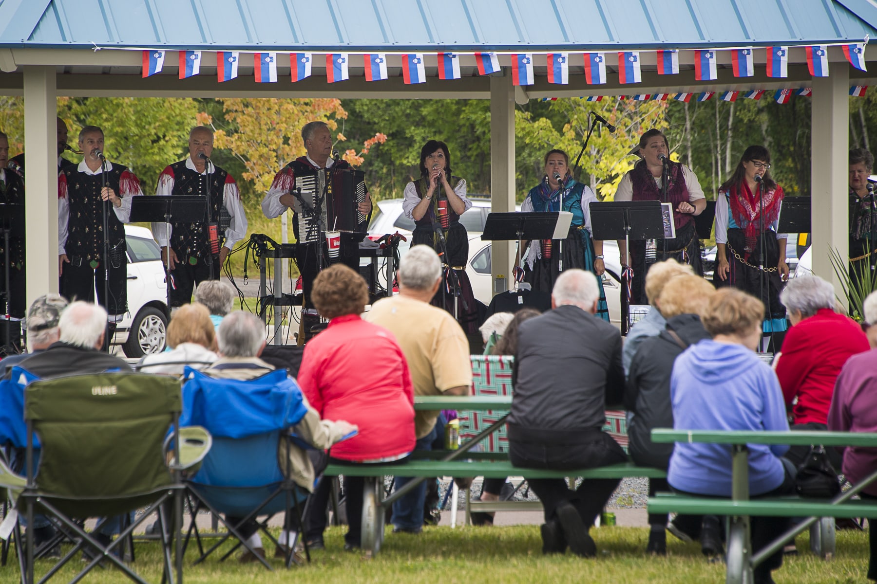A crowd sitting at picnic tables watches a group perform under a pavilion
