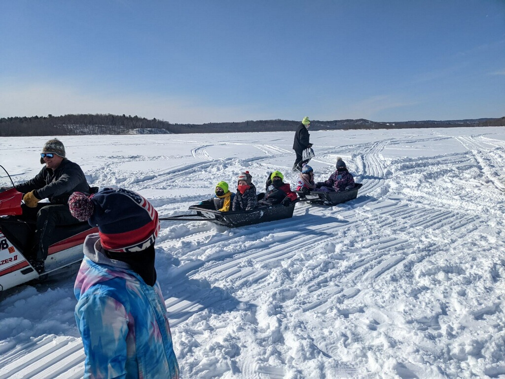 A group of children being pulled on two sleds by a snowmobile through the snow. Two people watch them
