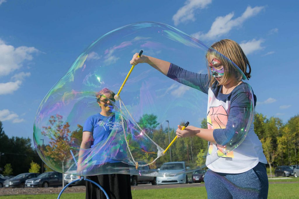 A young girl with face paint makes bubbles on a sunny day while a woman watches her and smiles