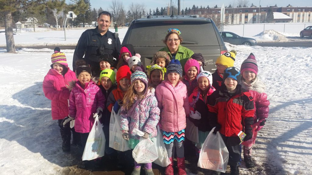 A group of children smile with a police man in front of a car in the winter