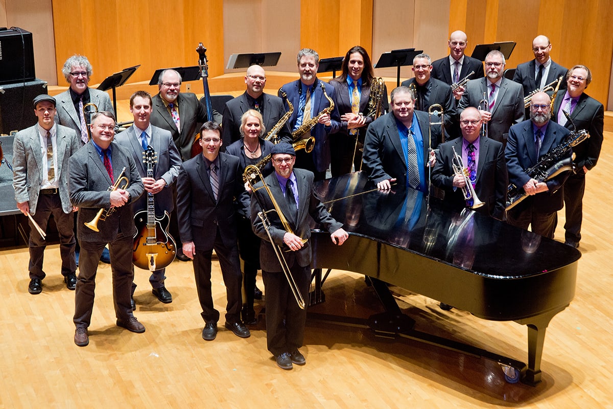 the Big Time Jazz Orchestra holding their instruments and standing near a grand piano in a concert space