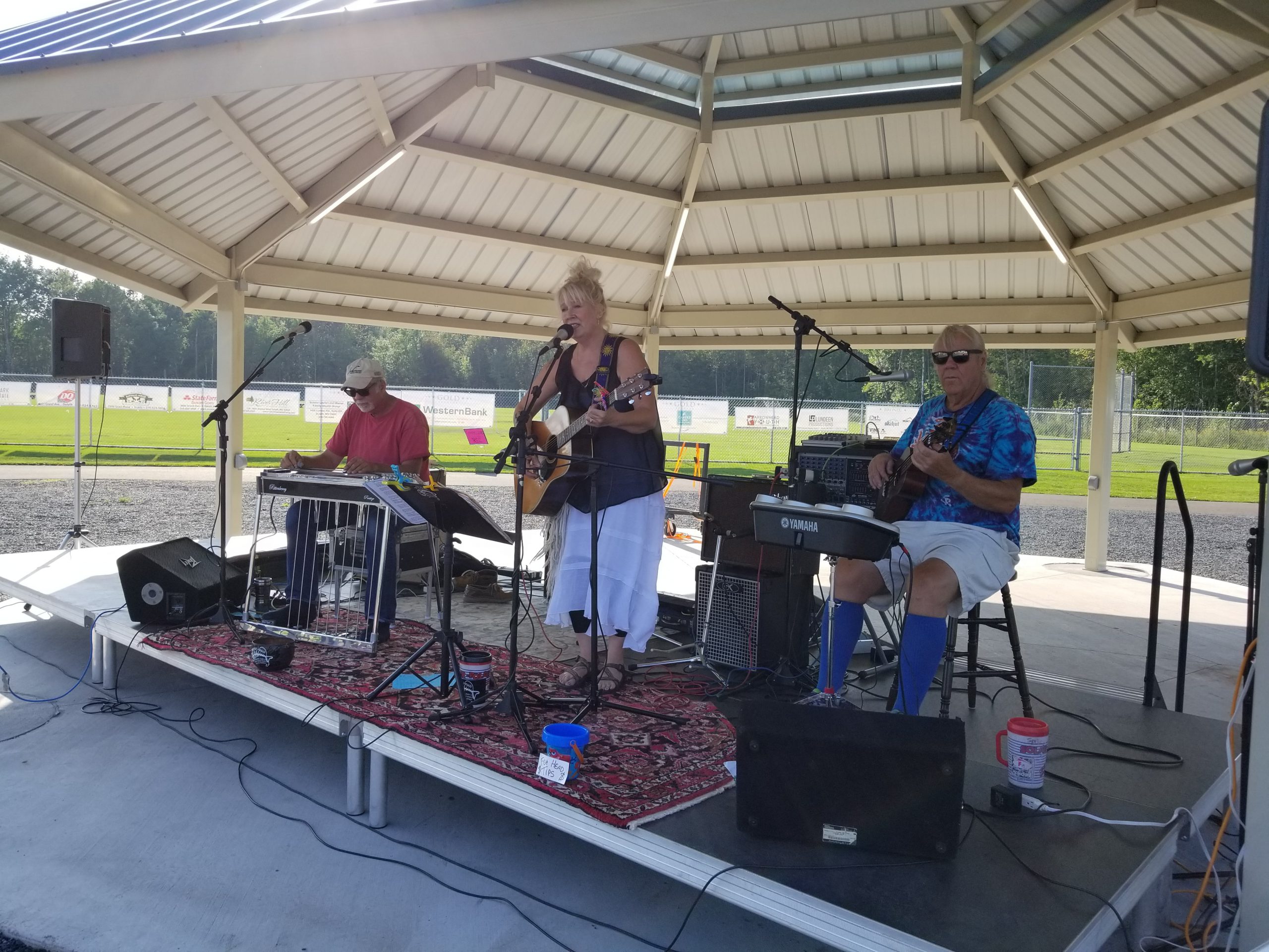 Two men playing keyboard and ukulele sit while a woman stands and sings at the Pavilion at the GND REC