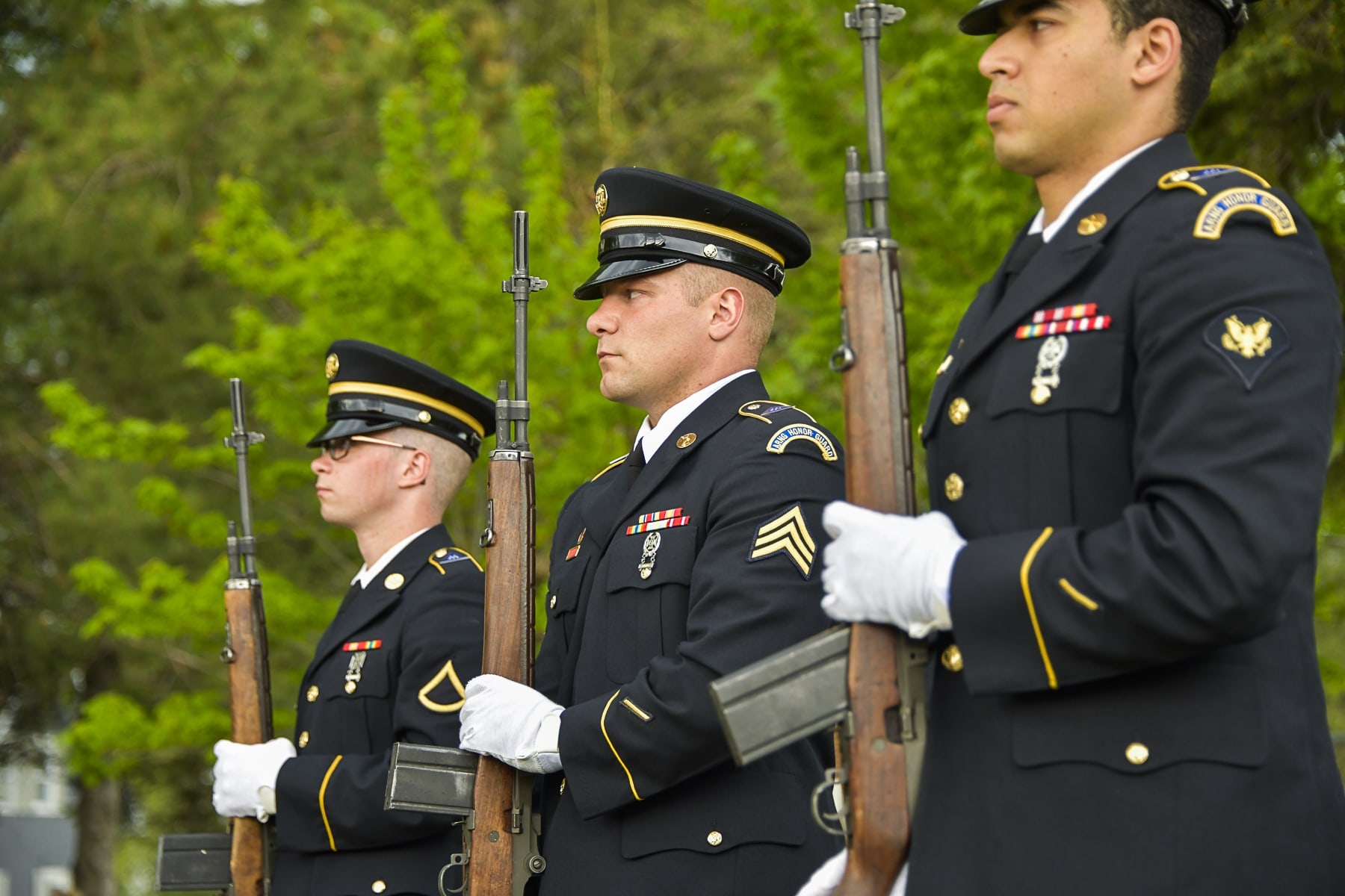 Three men in formal military uniforms hold rifles and stand at attention
