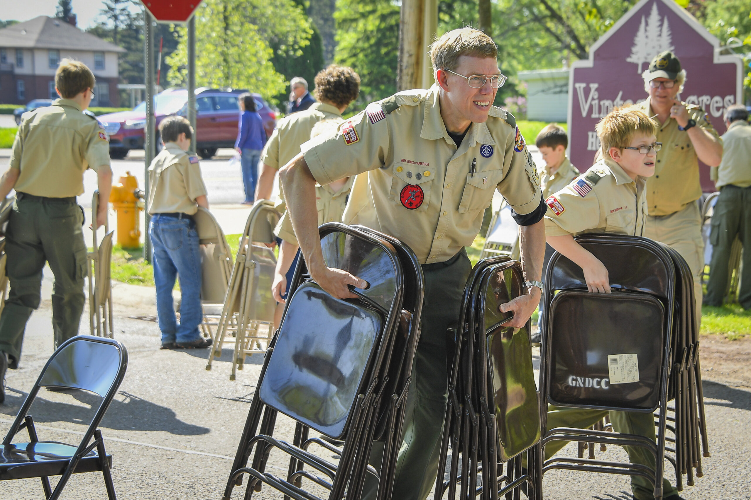 A man and a boy in boy scout uniforms hold metal folding chairs outside