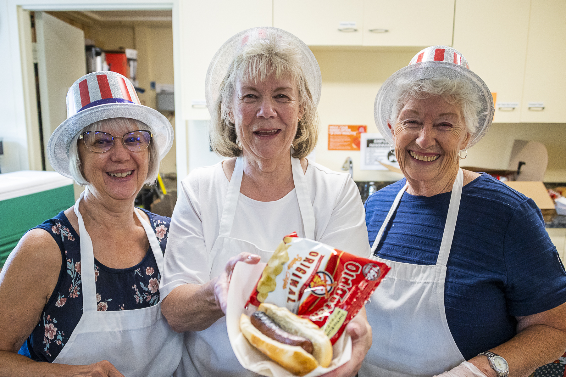 Three women wearing red, white, and blue hats smile while one holds up a plate with a hot dog and chips on it
