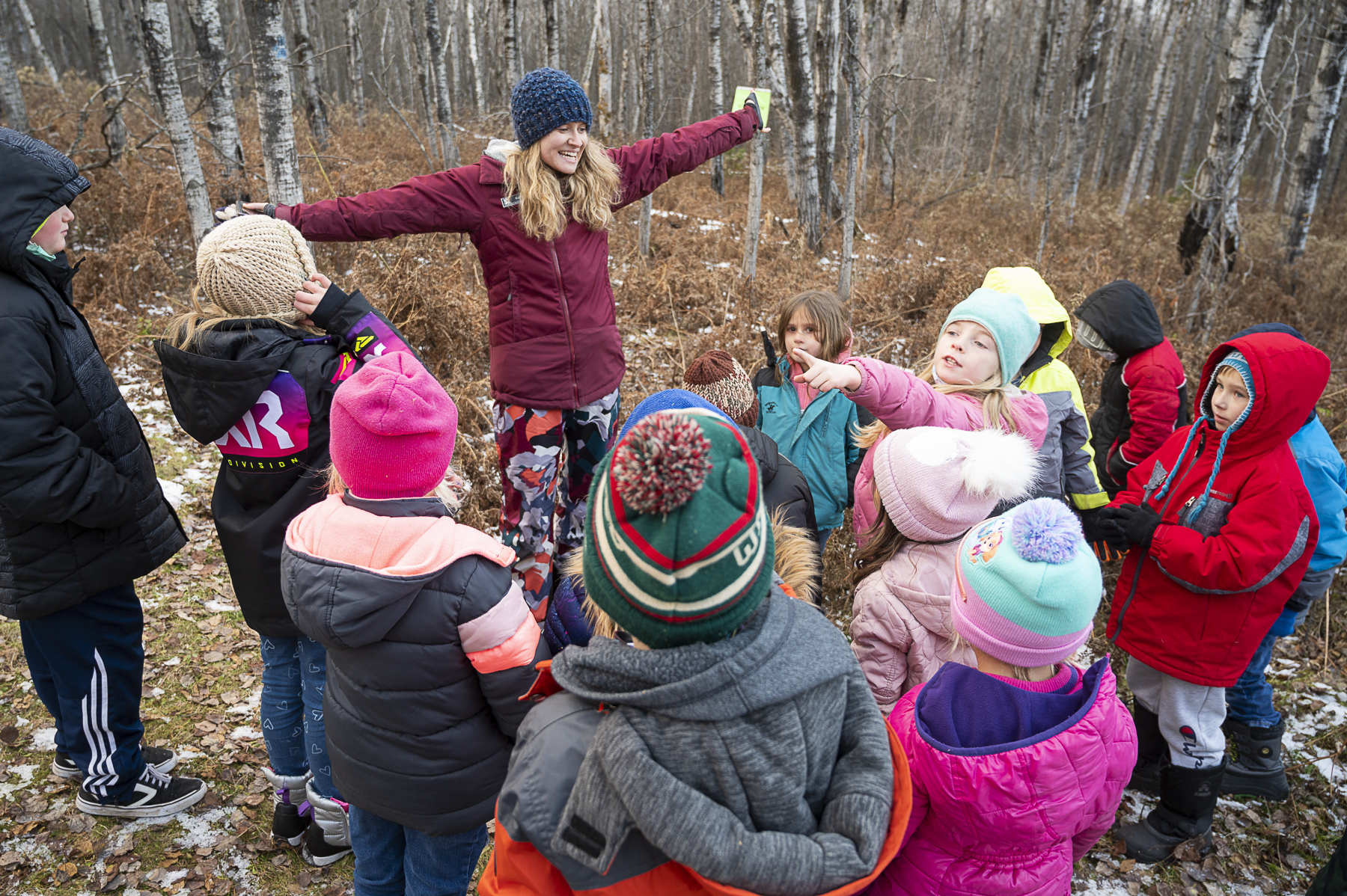 A woman with her arms open wide smiles at a group of children watching her in the woods at the GND REC