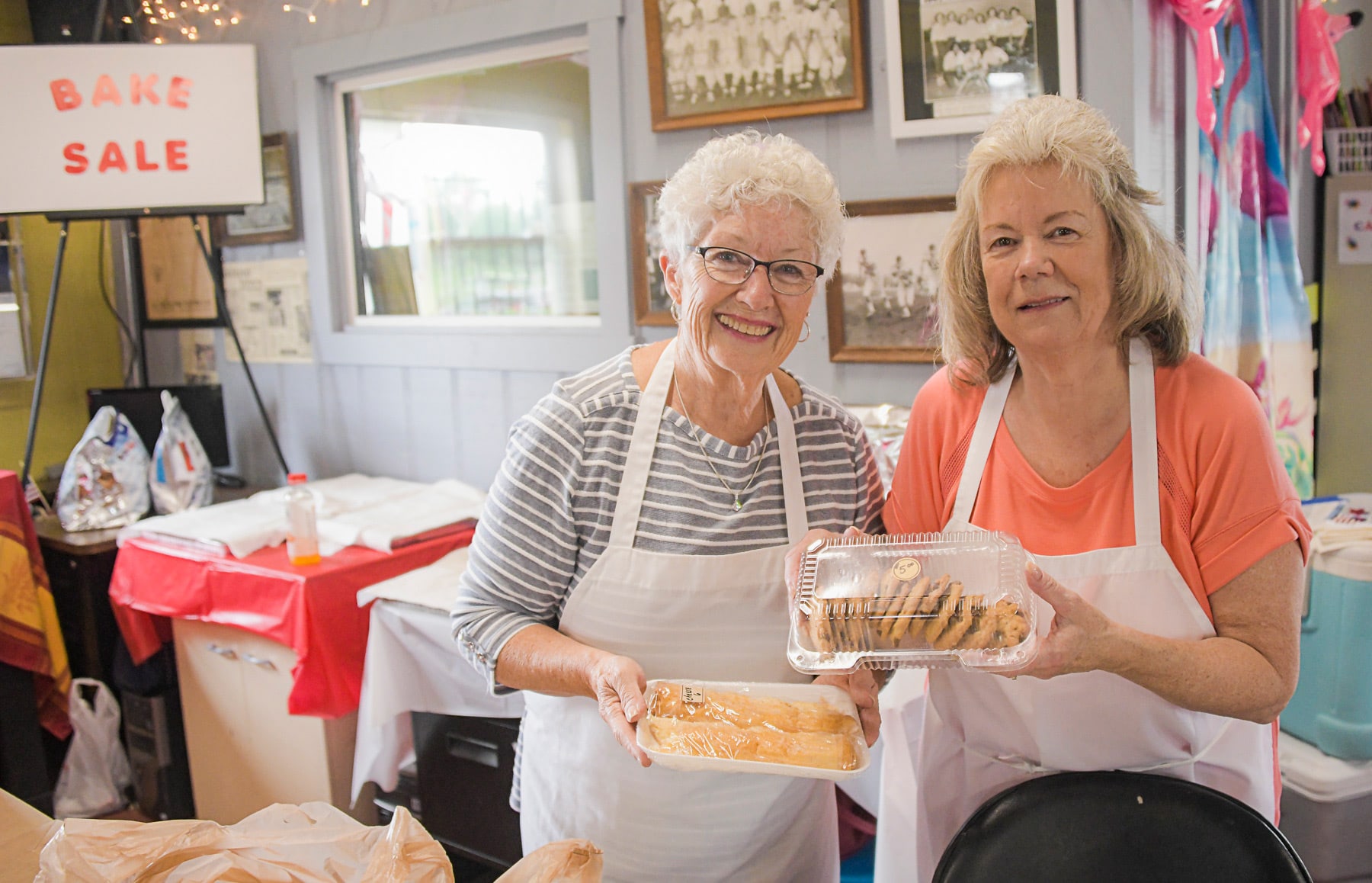 Two women hold up baked goods at a bake sale in the Community Building at the GND REC