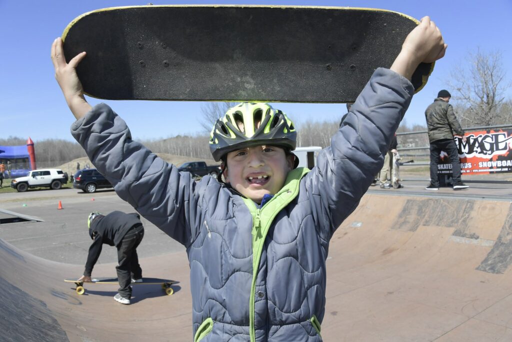 A young boy holds up a skateboard on a skate ramp