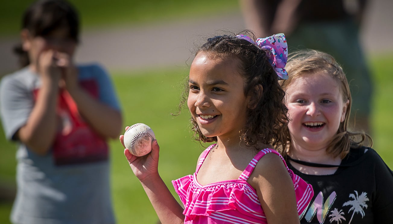 A young girl holds up a baseball while smiling while another girl smiles behind her