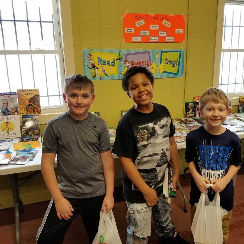 Three boys smile while holding bags and books in front of a book fair at the GND REC