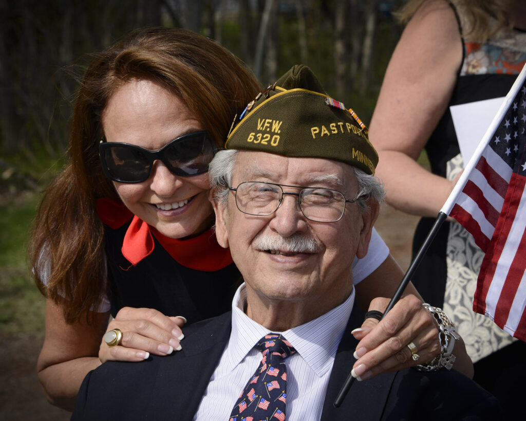 A woman holding an American Flag puts her hands on the shoulders of a man wearing a military hat while smiling