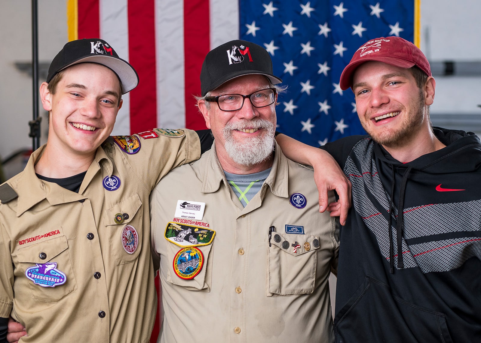 Two men in boy scout uniforms and one in a black sweatshirt pose in front of an American Flag